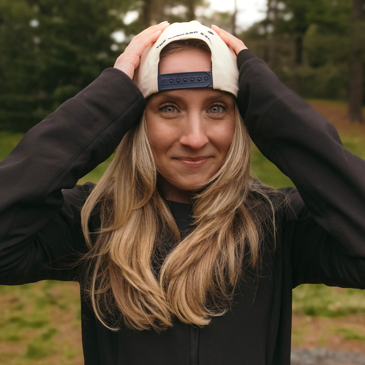 A woman with long blonde hair wearing a black zip-up jacket and a cream-colored baseball cap with a dark blue brand tab, posing outdoors with her hands on the cap in a forest setting.